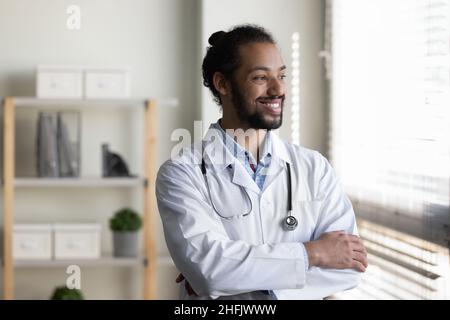 Happy dreamy young African American male doctor standing in office. Stock Photo