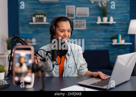 Woman vlogger with headphones looking at laptop and recording video. Influencer streaming online podcast on smartphone. Content creator in home studio using audio broadcasting equipment. Stock Photo