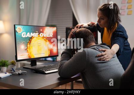 Couple feeling sad about losing video games play in front of computer. Woman comforting man about lost game online, having gaming equipment and headphones, playing for leisure. Stock Photo