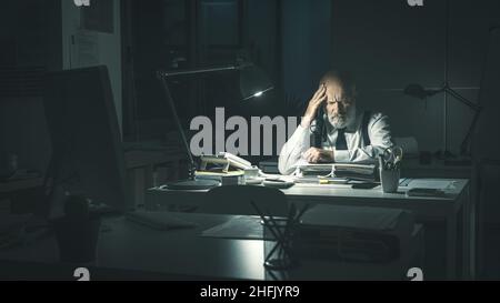 Stressed business executive sitting at desk and working overtime at night, he is having a bad headache Stock Photo