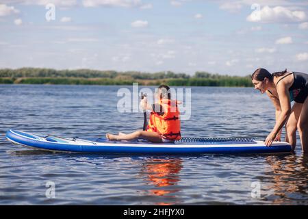 Mother helping little daughter to sup board with oar in hands on lake with green reeds and trees in background wearing vest life jacket. Active Stock Photo