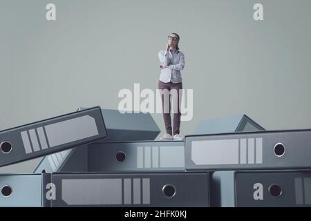 Businesswoman standing on a load of messy disorganized ring binders and thinking with hand on chin, data management concept Stock Photo