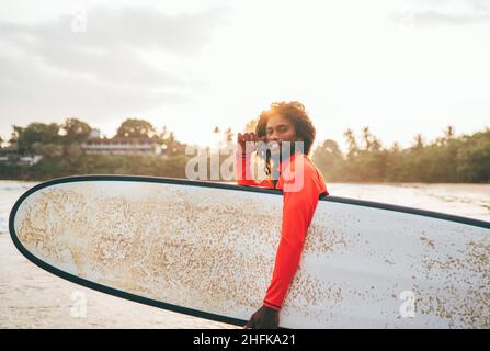 Portrait of black long-haired teen boy with a surfboard ready for surfing with sunset backlight. He walking into Indian ocean waves. Extreme water spo Stock Photo