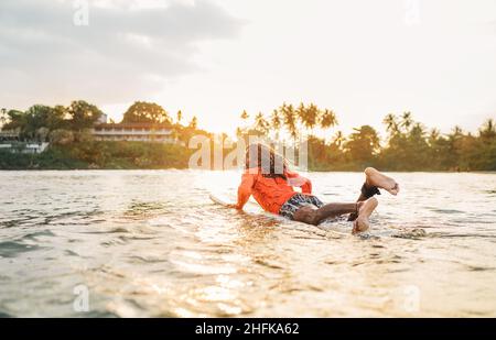 Black long-haired man paddling on long surfboard to the surfing spot in Indian ocean. Palm grove litted sunset rays in the background. Extreme water s Stock Photo