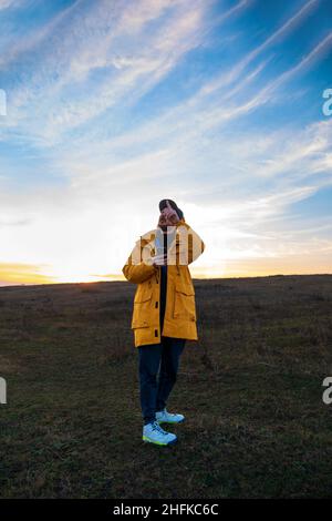 Traveler man in yellow coat with smartphone walking on a hill sunset sky side view. Authentic male tourist with gadget Stock Photo