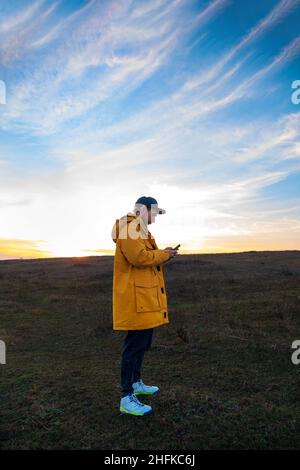 Traveler man in yellow coat with smartphone walking on a hill sunset sky side view. Authentic male tourist with gadget Stock Photo
