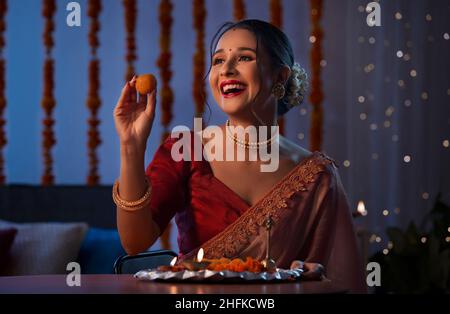 A beautiful woman holding a laddoo and smiling amidst diwali decoration,lights and pooja thali. Stock Photo