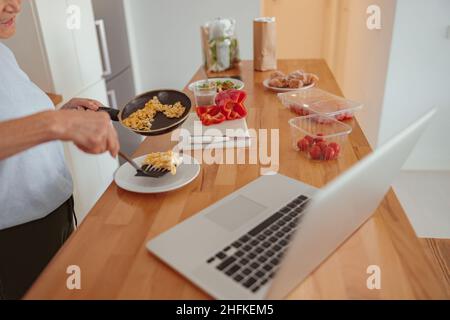 Pensioner female using laptop while cooking at kitchen Stock Photo