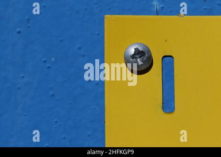 Macro close-up of a silver screw on a bright yellow metal plate against a textured blue background Stock Photo