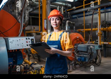 Concept of a industrial manufactory. A female smiling worker in uniform and helmet with a folder and a pen in her hand. In the background-boiler room. Stock Photo