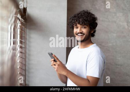 Happy indian man standing next to window in modern bathroom, using smartphone and smiling at camera Stock Photo