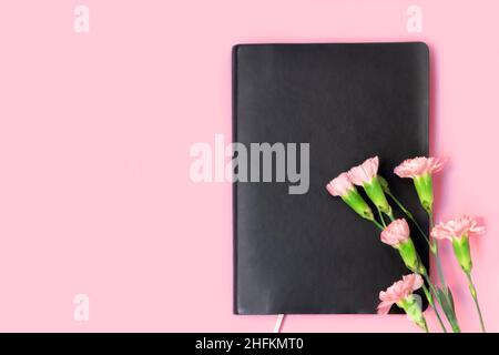 Above view of black empty notebook and carnation flowers on soft pink background with copy space. Stock Photo