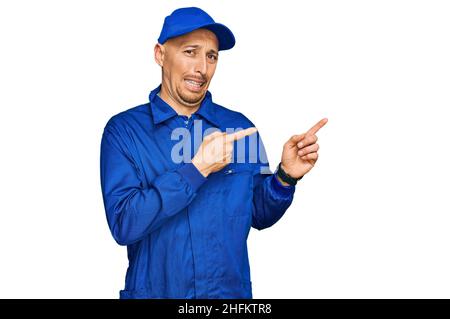 Bald man with beard wearing builder jumpsuit uniform pointing aside worried and nervous with both hands, concerned and surprised expression Stock Photo