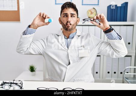 Young optician man holding glasses and contact lenses making fish face with mouth and squinting eyes, crazy and comical. Stock Photo