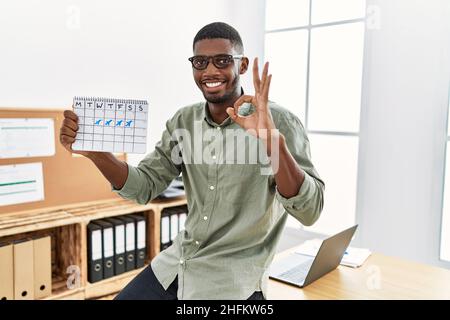 Young african american businessman holding travel calendar at the office doing ok sign with fingers, smiling friendly gesturing excellent symbol Stock Photo
