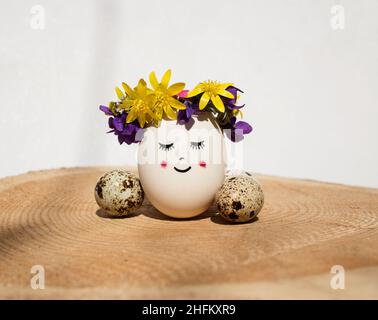 large egg with painted cute face with smile and closed eyes is decorated with wreath of fresh spring flowers. creative ideas for decor. happy Easter h Stock Photo