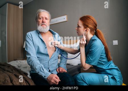 Female doctor holding stethoscope examining old senior male patient sitting on bed at home. Stock Photo