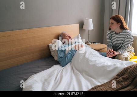 Sick senior aged male patient lying on bed in recovery room at hospital with granddaughter looking after Stock Photo