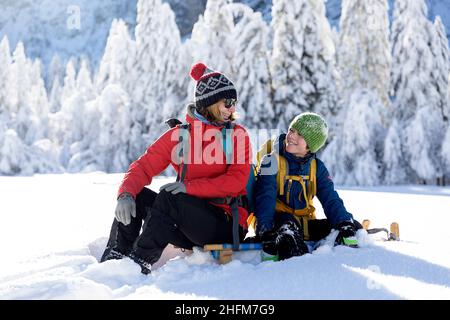 Mother and son sitting on sleds, looking at each other and smiling in fairytale winter landscape in Krnica valley, Slovenia Stock Photo