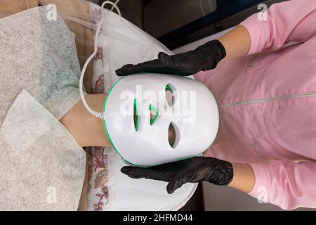 Woman lies in bed with led light therapy facial mask and relax. Led light mask for face skin treatment, frofessional cosmetology procedure. Stock Photo