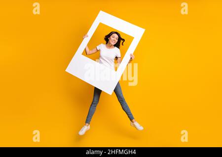 Full length body size view of lovely cheerful girl jumping having fun holding photo frame isolated over bright yellow color background Stock Photo