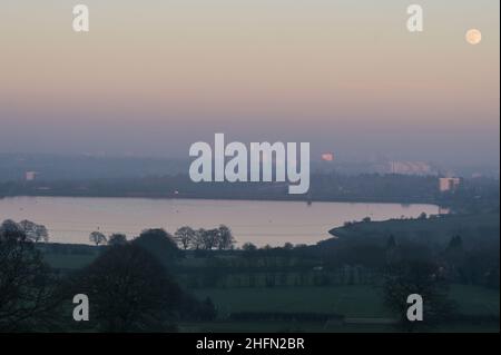 Frankley, Birmingham, UK. 17th Jan, 2022. The first full moon of 2022 called the Wolf Moon, rises above Frankley Reservoir in the foreground and Birmingham which is surrounded by thick haze blocking much of its view from the hills of Frankley, West Midlands. The large white building of Queen Elizabeth Hospital in Selly Oak is just visible below the moon. Pic by Credit: KStop Press Media/Alamy Live News Stock Photo