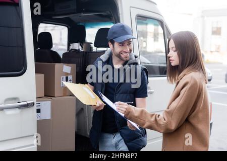 Smiling delivery man holding parcel near woman writing on clipboard and car outdoors Stock Photo
