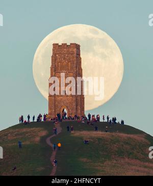 Glastonbury, Somerset, UK.  17th January 2022.  UK Weather.  The full Wolf Moon rises up from behind St Michael’s Tower on Glastonbury Tor in Somerset on a cold clear evening as people stand on top and watch it rise.  Picture Credit: Graham Hunt/Alamy Live News Stock Photo