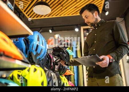 Bike shop manager checks helmet price information on tablet, seller makes an inventory in sports shop. Theme of small business selling bicycles Stock Photo