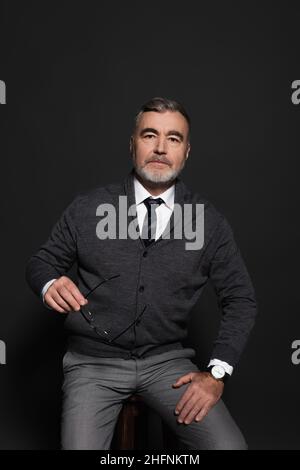 senior man in stylish clothes holding eyeglasses while sitting on dark grey Stock Photo
