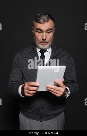 surprised senior man in jumper and tie looking at digital tablet isolated on dark grey Stock Photo