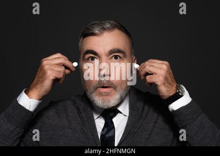 surprised senior man inserting earphones isolated on dark grey Stock Photo