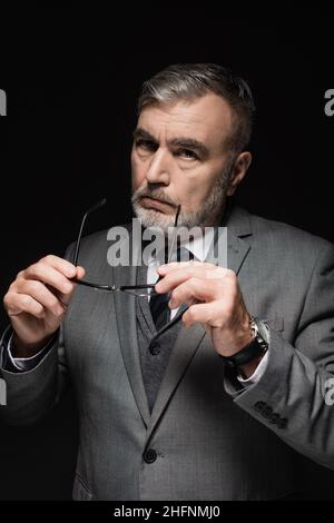 confident senior businessman in blazer holding eyeglasses and looking at camera isolated on black Stock Photo