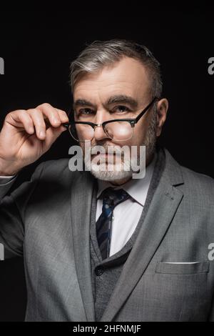 serious senior businessman adjusting eyeglasses while looking at camera isolated on black Stock Photo