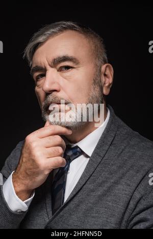 serious man in tie touching chin while looking at camera isolated on black Stock Photo