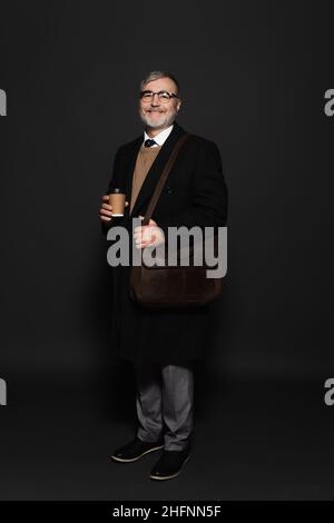 full length view of pleased senior man in black coat standing with leather bag on dark grey Stock Photo
