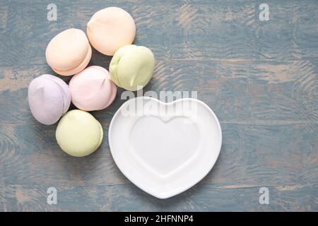 Round multi-colored marshmallows in a pile and a white plate in the shape of a heart on a blue wooden background, top view copy space. Delicious sweet Stock Photo