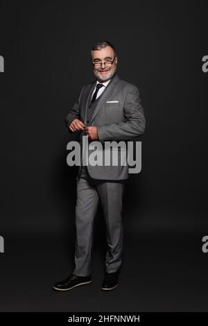 full length view of happy senior man in suit looking at camera on dark grey Stock Photo