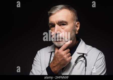 pensive bearded doctor looking at camera while touching chest isolated on black Stock Photo
