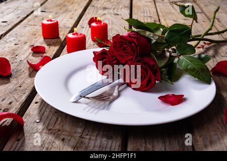 Fresh red roses with cutlery on plate by petals and burning candles at wooden table Stock Photo