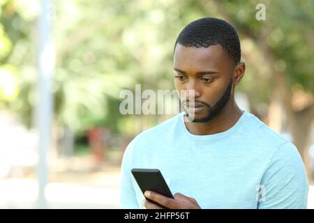 Worried man with black skin checking mobile phone walking in a park Stock Photo