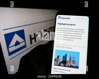 Person holding smartphone with webpage of German construction company Hochtief AG on screen in front of logo. Focus on center of phone display. Stock Photo