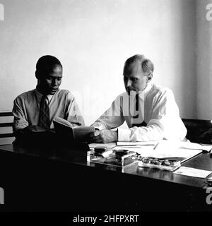 1950, historical, post-ww2 and a newly arrived immigrant sitting in an office beside a gentleman at a desk looking at a booklet, entitled, 'How To Live In Britain'. This offered advise on many topics, ranging from finding work and dealing with English landladies to queuing for a bus and the importance of drinking tea. Following the devastion of WW2, there was a shortage of manpower to rebuild the country and so immigration to Britain from the commonwealth countries was encouraged. An estimated one million people came to Britain from the West Indies ater the war. Stock Photo