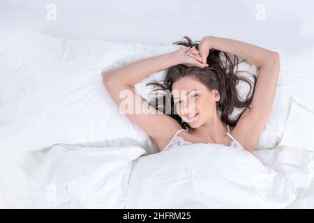 Wellness concept. Healthy lifestyle. A woman lies in bed and pulls her hands up after waking up. A young beautiful brunette woman wakes up in her bed Stock Photo