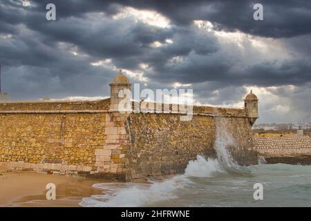 Waves break on the walls of the fortress Ponta da Bandeira, part of the maritime fortifications of Lagos, Algarve, Portugal. Dark, menacing clouds. Stock Photo