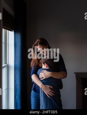 Happy mother and son embracing and smiling indoors beside a window. Stock Photo
