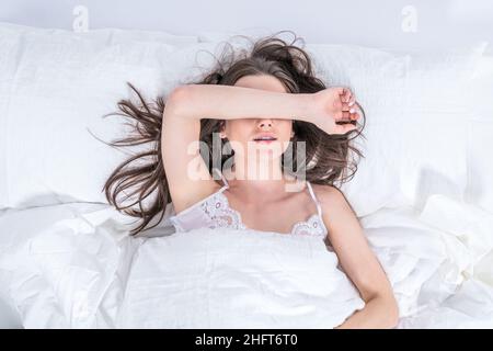 The brunette sleeps on a white bed. Bedroom. Good morning. Relaxation. A cute girl in pajamas lies on the bed, covering her eyes with her hand. Stock Photo