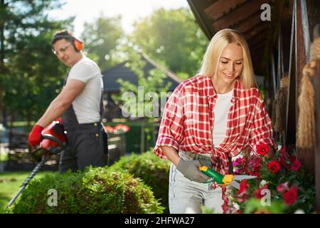 Attractive young woman with blond hair taking care of flower in pots while man trimming bushes on background. Two people working at garden during summer day.  Stock Photo