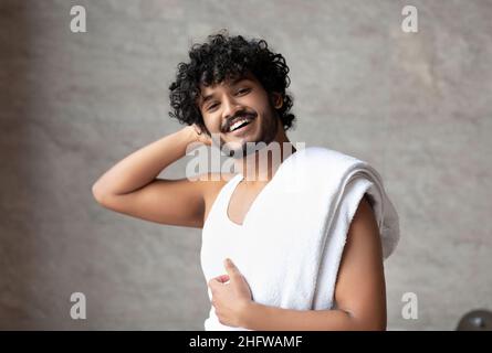 Male beauty care. Happy indian guy with towel on shoulder standing in bathroom and smiling at camera Stock Photo