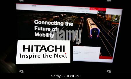 Person holding cellphone with logo of Japanese company K.K. Hitachi Seisakusho on screen in front of business webpage. Focus on phone display. Stock Photo
